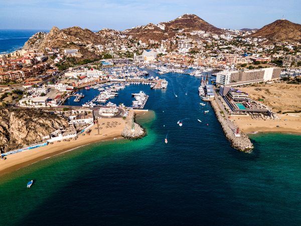 An aerial view of Cabo San Lucas Marina with boats docked, surrounded by mountains and sandy beaches, opening into the blue waters