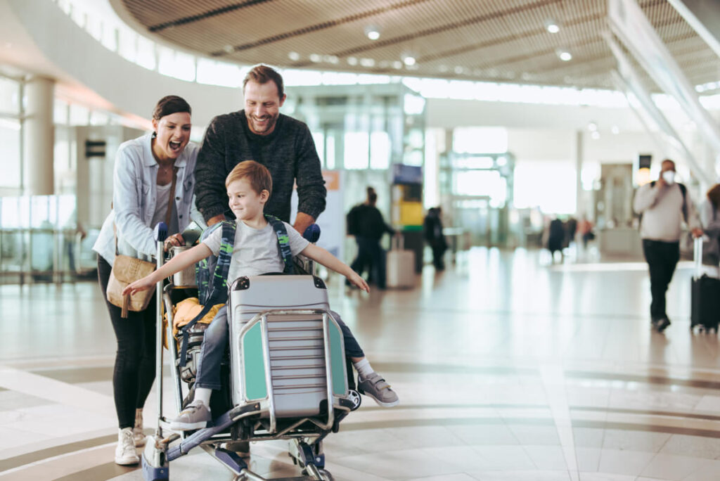 Couple happily pushing the trolley with their son at airport. Child enjoying sitting on luggage trolley
