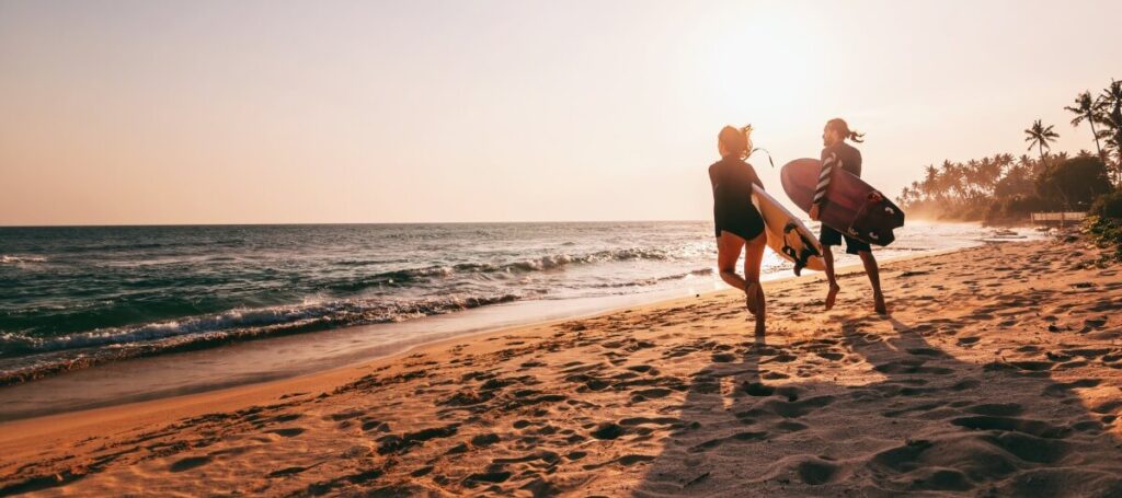 Young beautiful couple walking along the sandy beach near the ocean at sunset with surfboards, outdoor activities